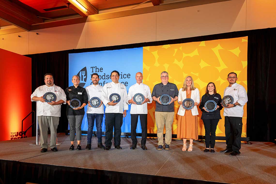 Nine people standing on a stage holding plates, smiling. The backdrop features a blue and orange display with the text "The Foodservice Conference." The group appears to be diverse, including men and women, dressed in business casual and chef attire.