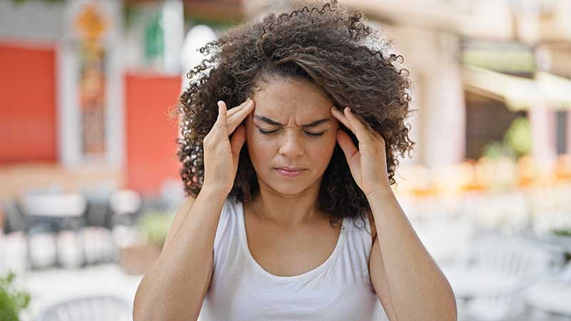 A person with curly hair holds their temples with closed eyes, appearing uncomfortable, in an outdoor setting.