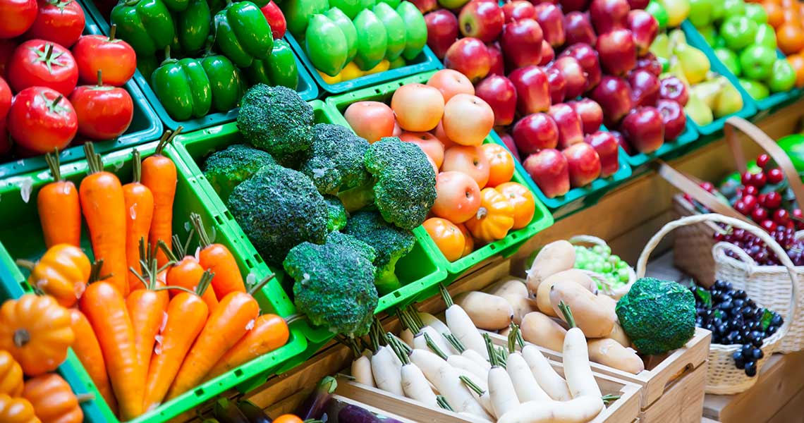 Colorful market display of various fruits and vegetables including carrots, pumpkins, broccoli, apples, and grapes.