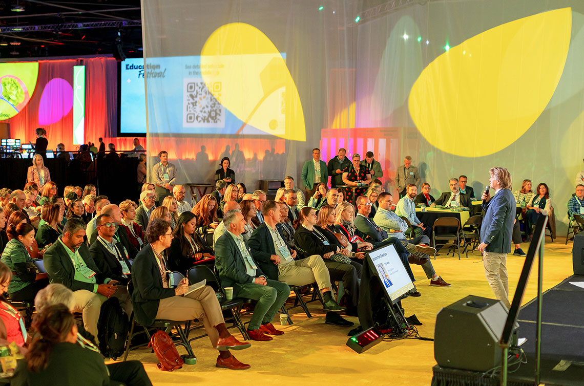 A speaker addresses an audience during the educational festival at the Global Produce and Floral Show, with colorful abstract projections in the background.